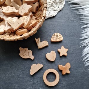 Op de afbeelding: Een set houten baby-bijtjes in verschillende vormen, waaronder een wolk, een hart, een ster, een regenboog, een blad, een vlinder, een kat en een ring. De bijtjes zijn van natuurlijke houtkleur en hebben een gladde, gepolijste afwerking.