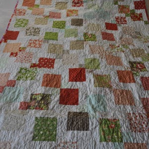 May include: A handmade quilt featuring a patchwork design with squares in shades of red, orange, green, and brown on a white background. The quilt has a pink border and is likely made of cotton fabric.