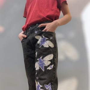 May include: Black denim jeans featuring a series of white and purple cicada insect graphics printed along the front leg. The jeans have a standard five-pocket design with a button closure and are paired with a plain red short-sleeved t-shirt.