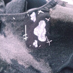 Peut inclure: Une veste en jean noire avec un patch noir cousu sur l'&eacute;paule gauche. Le patch a des coutures blanches et des &eacute;claboussures de peinture blanche qui ressemblent &agrave; des nuages.