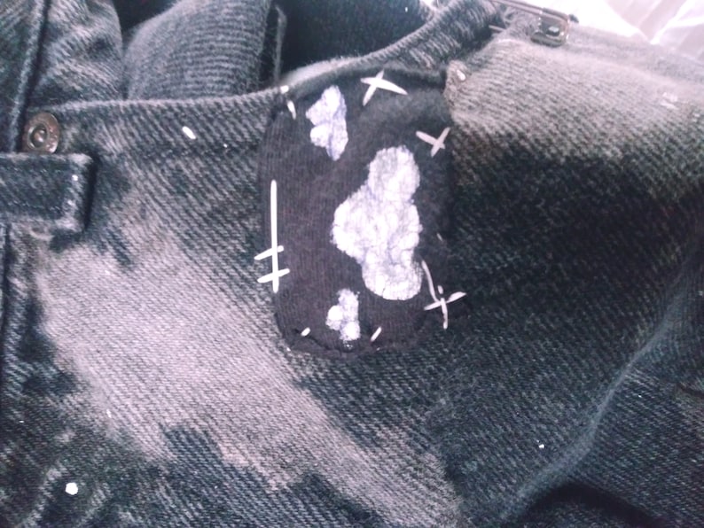 Peut inclure: Une veste en jean noire avec un patch noir cousu sur l'&eacute;paule gauche. Le patch a des coutures blanches et des &eacute;claboussures de peinture blanche qui ressemblent &agrave; des nuages.