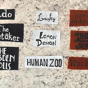 Peut inclure: Une collection de neuf écussons rectangulaires en tissu avec du texte peint à la main. Les écussons portent les noms Ado, Laufey, danny gonzalez, The Caretaker, LEMON DEMON, THAT HANDSOME DEVIL, THE DRESDEN DOLLS, HUMAN ZOO et WILL WOOD en encre noire ou blanche.