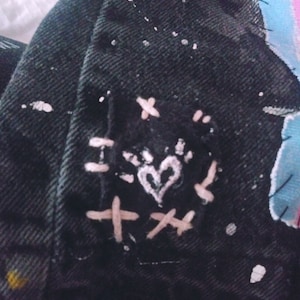 Peut inclure: Veste en jean noire avec des &eacute;claboussures de peinture blanche et un patch brod&eacute; en forme de c&oelig;ur rose. Le patch est entour&eacute; de coutures blanches en forme de X.