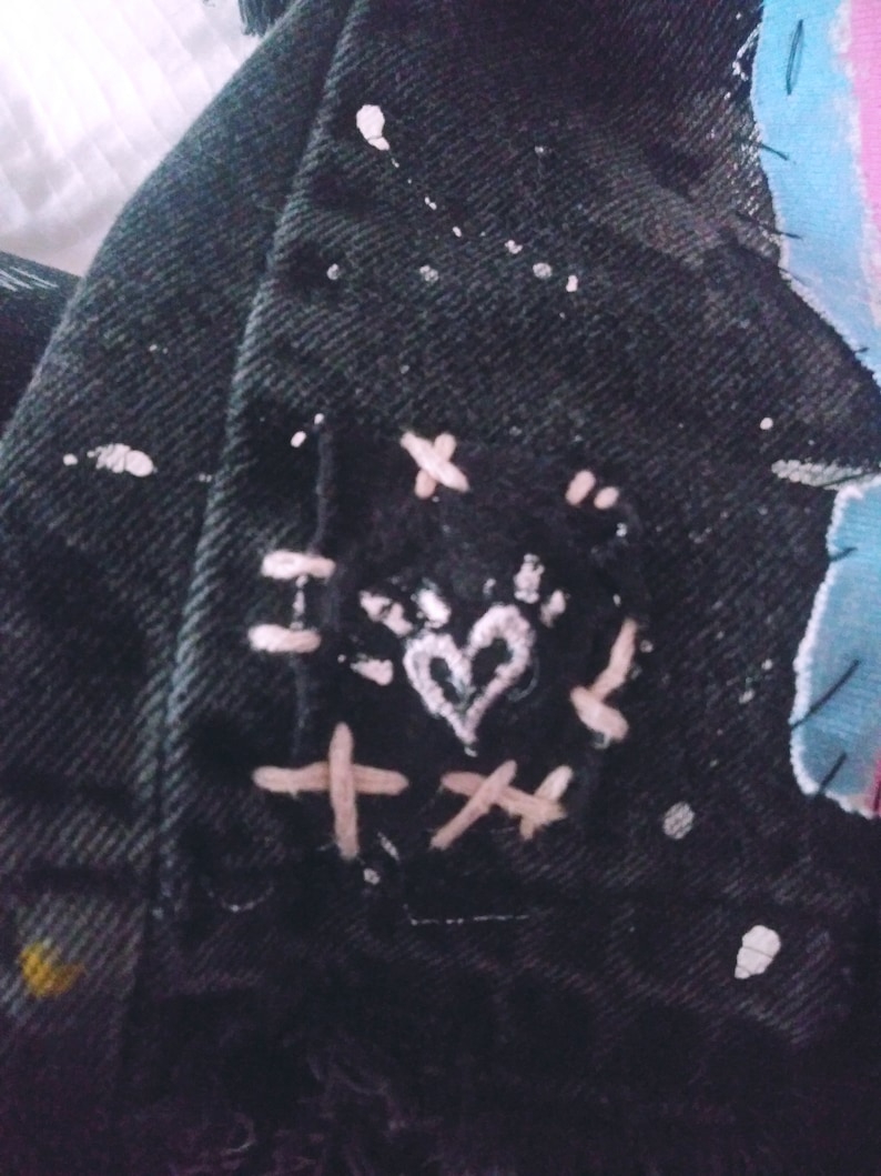 Peut inclure: Veste en jean noire avec des &eacute;claboussures de peinture blanche et un patch brod&eacute; en forme de c&oelig;ur rose. Le patch est entour&eacute; de coutures blanches en forme de X.