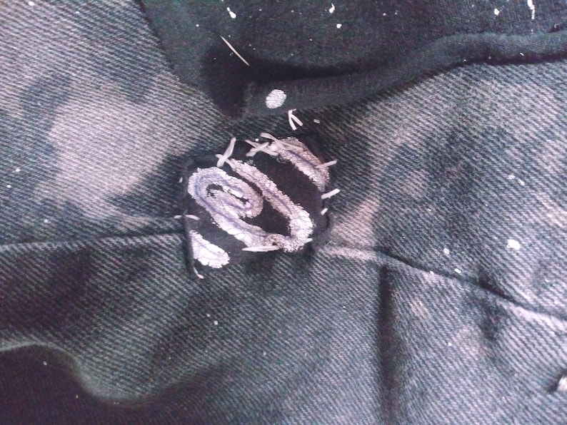 Peut inclure: Un gros plan sur un patch en tissu noir avec un motif en spirale blanc, cousu sur un tissu denim fonc&eacute;.