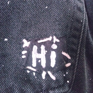 Peut inclure: Une veste en jean noire avec un patch brod&eacute; blanc qui dit "HI" dessus.