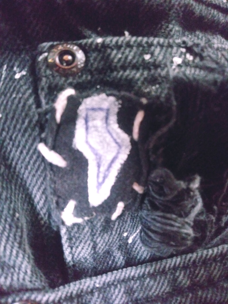 Peut inclure: Gros plan sur une poche en jean noir avec un patch en forme d'&eacute;clair blanc et bleu. La poche a un bouton en m&eacute;tal et est partiellement d&eacute;chir&eacute;e.