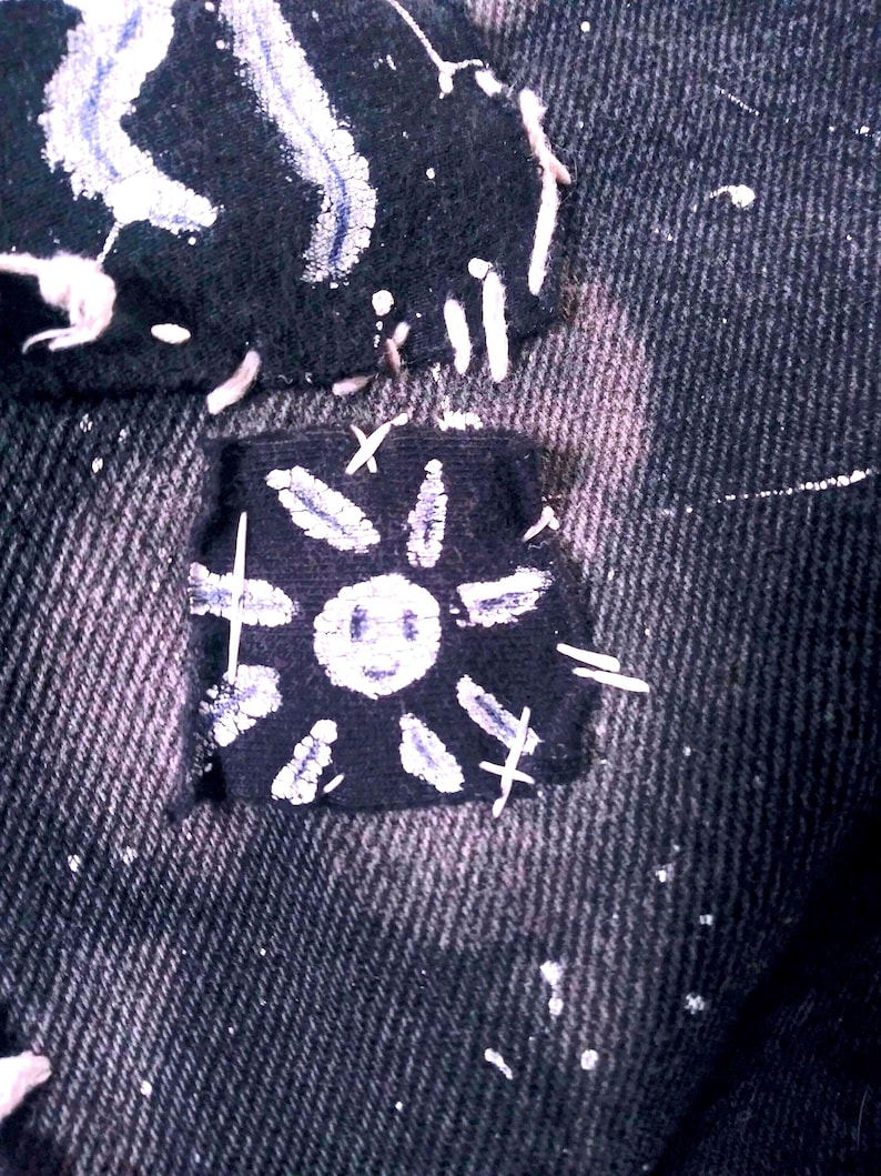 Peut inclure: Une veste en jean noire avec un patch en forme de soleil blanc cousu sur le devant. Le soleil a un visage souriant et des rayons.