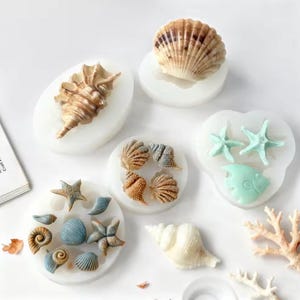 May include: A collection of white silicone molds for casting various sea-themed shapes, including detailed conch shells, spiral shells, starfish, and a small fish. Several finished resin or clay figures in shades of blue, brown, and white are displayed.