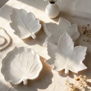 May include: A set of four white ceramic dishes shaped like various leaves, featuring detailed embossed veins on the surface. The collection includes three distinct leaf designs with smooth, glossy finishes, arranged together on a flat surface.