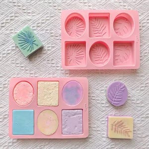 May include: Two rectangular pink silicone molds with six cavities each, featuring various geometric shapes and botanical leaf patterns. Several finished soap bars in pastel colors, including blue, purple, and yellow, are displayed alongside.