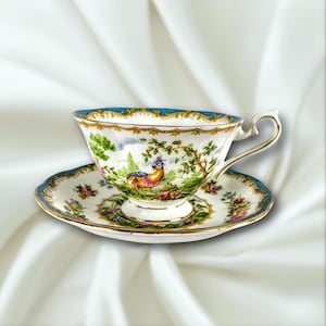 May include: A vintage teacup and saucer set. The white porcelain cup and saucer feature a detailed landscape scene with a colorful bird, trees, and floral accents. The rims are trimmed with gold and a teal blue border.
