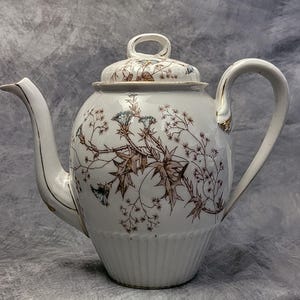 May include: White ceramic teapot with a lid, featuring a floral design in brown, blue, and gold. The teapot has a curved spout and handle, with a ribbed lower section. The lid has a small loop handle. A decorative piece for serving tea.