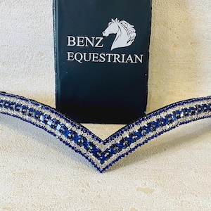 以下が含まれることがあります： 青とシルバーの馬術用額革、V字型デザイン。 額革は青とシルバーのクリスタルで飾られています。 背景には「BENZ EQUESTRIAN」の文字と馬の頭のロゴが入った黒いカードがあります。