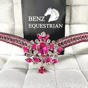 Puede incluir: Primer plano de una frontalera ecuestre rosa y plateada. La frontalera presenta un grupo central de cristales rosas y transparentes, con un colgante en forma de lágrima. La marca "BENZ EQUESTRIAN" es visible en una etiqueta negra.