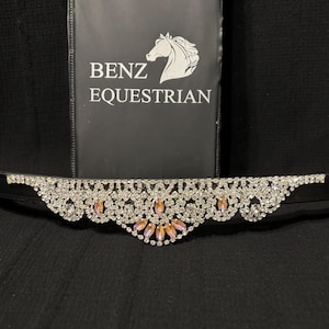 Puede incluir: Un frontal de cabezada de cuero negro con un diseño de cristal y strass rosa. El frontal presenta un patrón delicado e intrincado con un grupo central de strass rosa. El frontal está adornado con el texto "Benz Equestrian".