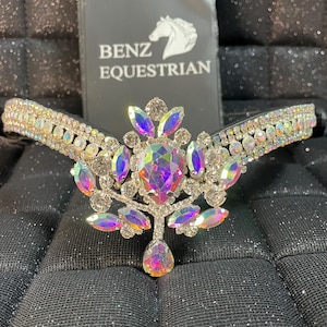 Puede incluir: Una frontalera ecuestre plateada adornada con cristales iridiscentes y un colgante central en forma de lágrima. La frontalera presenta un diseño floral y el logotipo dice "BENZ EQUESTRIAN". El fondo es una superficie negra texturizada.