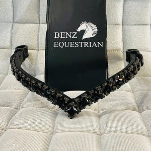 Puede incluir: Una cabezada ecuestre negra con un diseño en forma de V, adornada con piedras preciosas oscuras. La cabezada está colocada frente a una etiqueta negra de Benz Equestrian con un logotipo de cabeza de caballo.