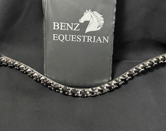 May include: A curved browband featuring a row of faceted black crystals set in metal, positioned in front of a black rectangular case with the white text BENZ EQUESTRIAN and a white horse head logo printed on the front surface of the packaging.