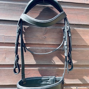 Op de afbeelding: Een zwart lederen paardenhoofdstel met een gevoerd kopstuk, een gebogen frontriem en een brede, gevoerde neusriem. Het hoofdstel is voorzien van verstelbare bakstukken met zilverkleurige metalen gespen en lederen keepers.