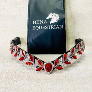 Puede incluir: Un frontal de caballo de cuero negro con un diseño de cristal plateado y rojo. El frontal presenta un diseño en V con cristales rojos en el centro y cristales plateados en los lados. El frontal está decorado con un patrón de hojas. El frontal está sobre un fondo negro con el texto "Benz Equestrian".