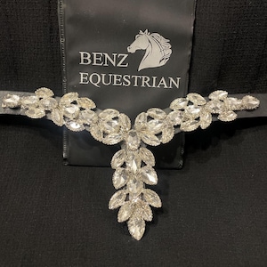 Puede incluir: Una frontalera ecuestre negra adornada con cristales transparentes. El diseño central presenta una cascada de cristales, con arreglos florales y en forma de hojas. El logotipo de Benz Equestrian es visible en el fondo.