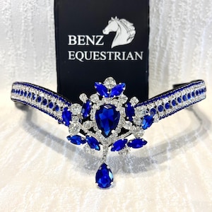 Puede incluir: Un frontal de caballo plateado y azul con un gran colgante de cristal azul en forma de lágrima y cristales azules más pequeños. El frontal está decorado con pedrería plateada y el texto "Benz Equestrian" es visible en el fondo.