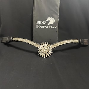 Puede incluir: Cabezada de cuero negro para caballo con un diseño floral plateado central y una fila de pedrería transparente. La cabezada es curva y tiene una etiqueta Benz Equestrian en el fondo.