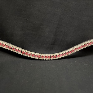 May include: A curved, decorative browband for a horse bridle. The browband is black with a row of small, bright pink gemstones set between two rows of clear, sparkling rhinestones. The background is solid black.
