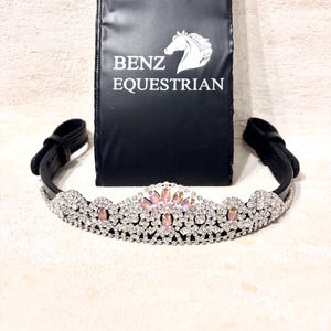 Peut inclure: Un frontal de cheval en cuir noir avec un design de couronne en cristal argenté et rose. Le frontal est décoré d'une grande couronne en cristal ornée de cristaux roses et transparents. Le frontal est emballé dans un sac en velours noir avec le texte "Benz Equestrian" imprimé dessus.