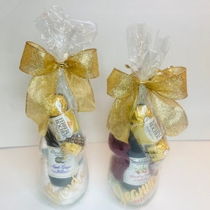 Op de afbeelding: Twee glazen flessen met gouden strikken en gepersonaliseerde namen "Mandie" en "Jeanne". Elke fles bevat een mini fles mousserende wijn, een mini fles likeur en een paar Ferrero Rocher chocolaatjes.