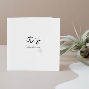 May include: A white card with the words "it's natural to cry" in black script. A small, light blue teardrop graphic is below the text. The card is on a neutral surface with a plant in the background.