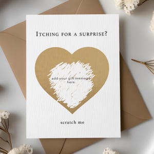 May include: A white card with the text "Itching for a Surprise?" and "scratch me". A gold heart shape is in the center with a scratch-off area. The card is on a brown envelope.