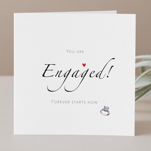 May include: White greeting card with the words "You are Engaged!" in elegant script, a small red heart, and the phrase "Forever Starts Now." A diamond engagement ring is pictured at the bottom.