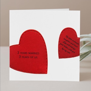 May include: A white greeting card featuring two red heart-shaped cutouts with stitched edges. The larger heart displays the text 3 YEARS MARRIED 3 YEARS OF US. The smaller heart displays the text Of little moments, laughter and trust. Life will always feels better with you by my side.