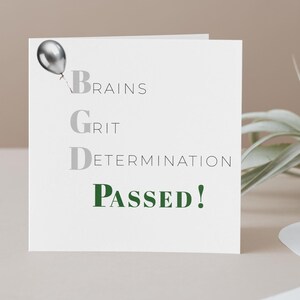 May include: A white greeting card with the words "Brains, Grit, Determination, Passed!" in black and green text. A silver balloon graphic is in the upper left corner. The card is square and has a minimalist design.