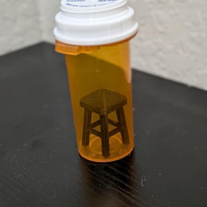 May include: An orange prescription pill bottle with a white cap contains a small, dark brown wooden stool. The stool is a miniature replica with four legs and a square seat. The bottle is on a black surface.