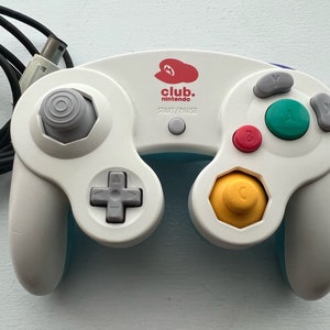 May include: White Nintendo GameCube controller with a blue and yellow faceplate. The controller has a red and white "club. nintendo" logo on the top. The controller has a gray start/pause button and a gray analog stick.