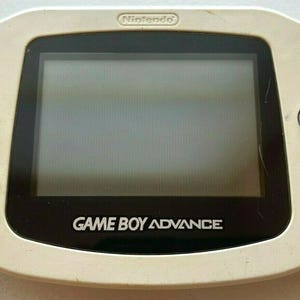 May include: A white Game Boy Advance handheld gaming console with a black screen and buttons. The console has a silver Nintendo logo on the top and the words "GAME BOY ADVANCE" printed on the bottom.
