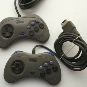 May include: Two gray Sega controllers with black buttons and a blue start button. The controllers have black cords and are connected to a Sega Genesis console.