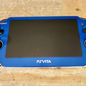 May include: A blue Sony PlayStation Vita handheld gaming console. The device has a black screen, a directional pad, and action buttons. The text "PS VITA" is printed on the bottom of the console.