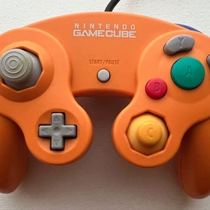 May include: An orange Nintendo GameCube controller with gray buttons and a white start/pause button. The controller has a black cord attached to the top.