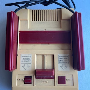 May include: A beige and red Nintendo Family Computer console with a black power cord. The console has a red power button, a red eject button, and a red reset button. The console has Japanese text on it.