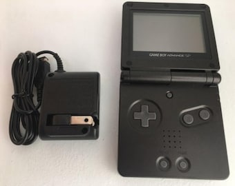 Könnte beinhalten: Eine schwarze Game Boy Advance SP Handheld-Spielkonsole mit einem schwarzen Netzteil und Kabel. Die Konsole hat einen Bildschirm, ein Steuerkreuz und Tasten. Der Text "GAME BOY ADVANCE SP" ist auf der Konsole aufgedruckt.