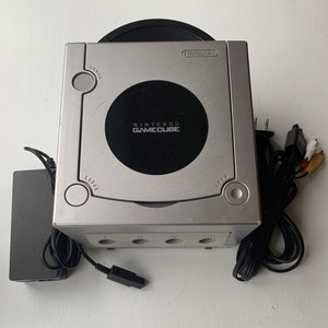 May include: A silver Nintendo GameCube console with a black disc drive and four controller ports. The console is connected to a black power cord and a black AV cable.