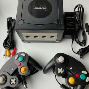 May include: A black Nintendo GameCube console with two black controllers, a power adapter, and an AV cable. The console has a circular disc drive and four memory card slots.