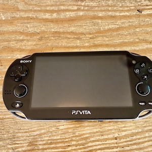 May include: Black Sony PS Vita handheld game console. The device has a large screen, a directional pad, and buttons on the left side, with buttons and a joystick on the right. The PS Vita logo is visible on the bottom.
