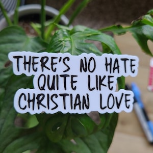 Puede incluir: Pegatina blanca con texto negro que dice "There's no hate quite like Christian love." La pegatina está en una planta verde.