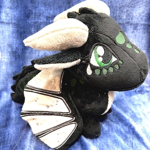 May include: A plush dragon toy featuring soft black fabric with white accents on the horns and wings. The creature has large embroidered eyes with green irises and white highlights, along with small green embroidered spots on its face and back.