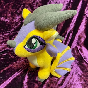 May include: A soft toy dragon in yellow, purple, and grey. The dragon features large eyes with green irises and purple outlines. It has wings with yellow and purple details, and grey horns. The toy is made of soft fabric.